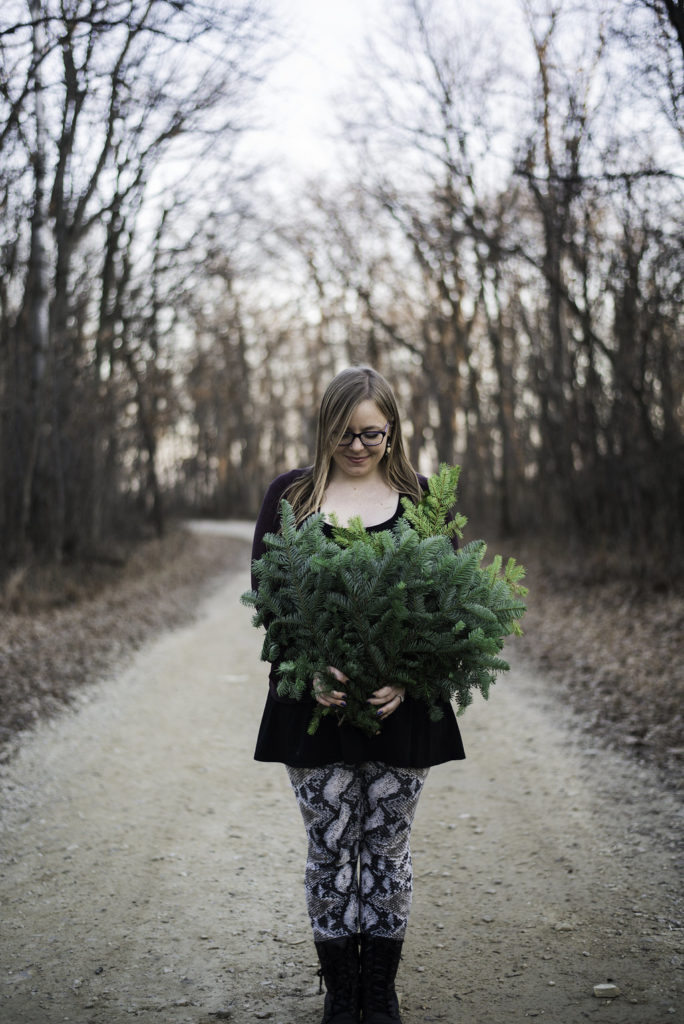 White woman holding evergreen branches in the middle of a dirt road in the winter. 